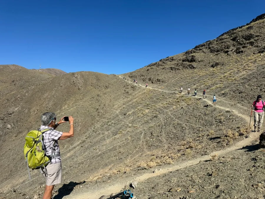 A hiker photographing a rocky trail winding across a dry mountain slope