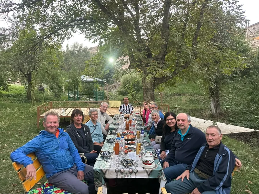 Travelers sharing a long outdoor meal beneath leafy garden trees
