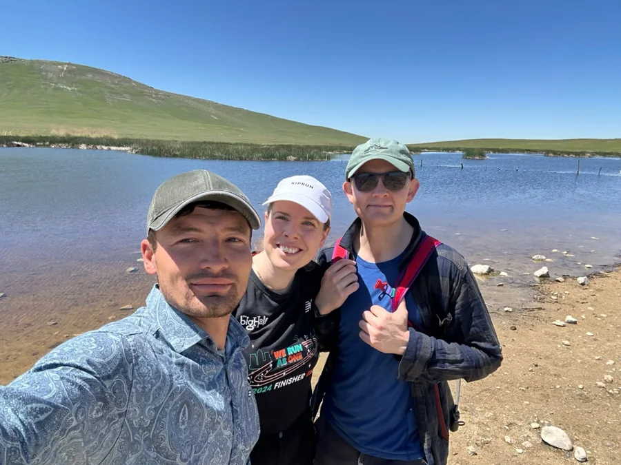 Three travelers taking a photograph together beside a blue lake