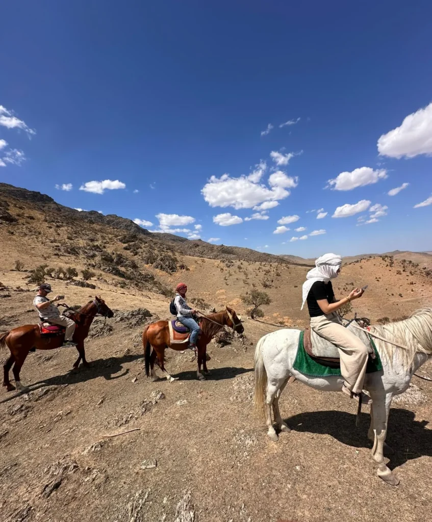 Three travelers ride horses across a dry mountain trail beneath a deep blue sky