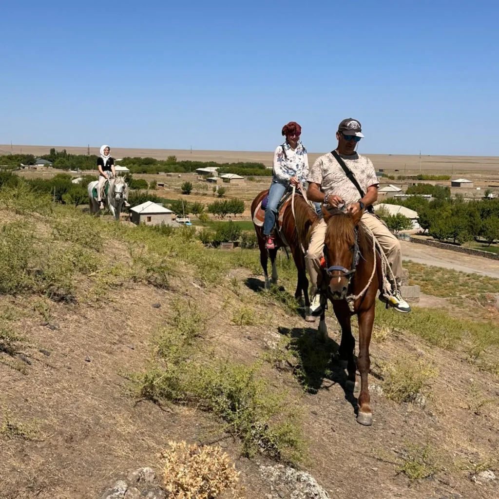 Three horseback riders descend a hillside above a green village in the Nuratau foothills of Uzbekistan