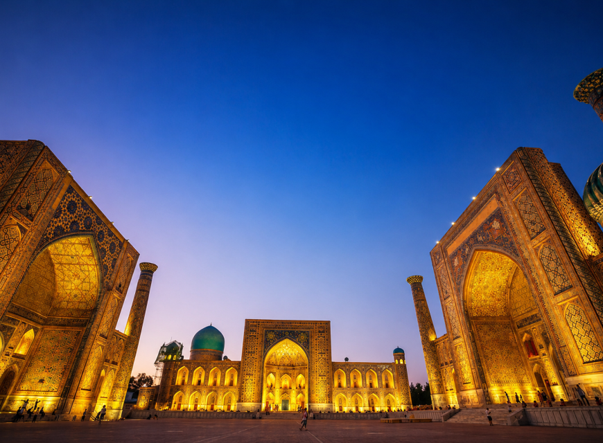 Wide-angle dusk view of Registan Square in Samarkand, Uzbekistan, featuring three grand madrasahs illuminated with warm night lighting under a deep blue sky
