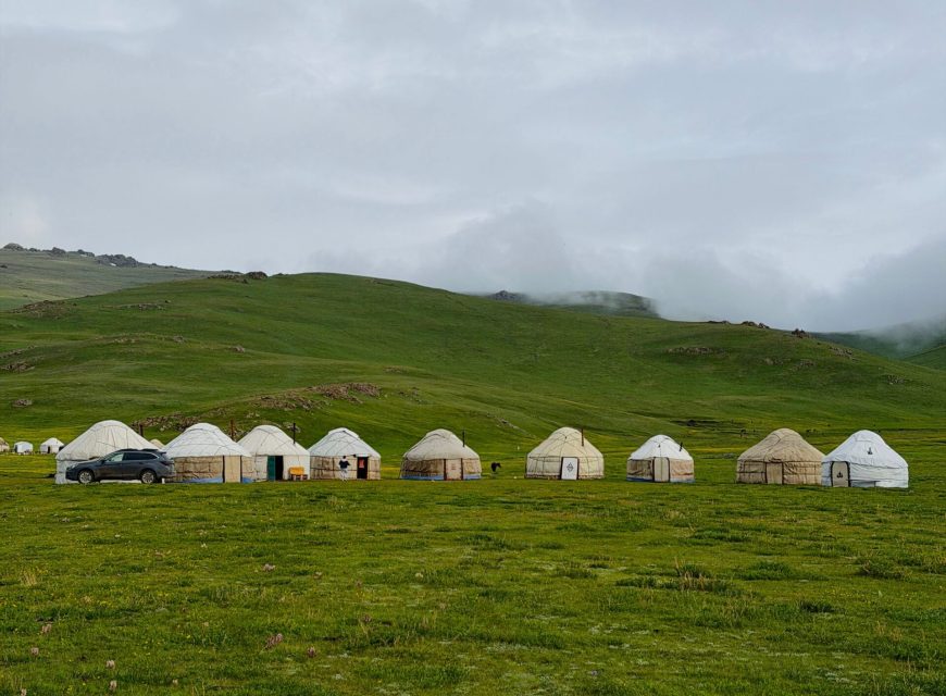 Vertical view of a line of white nomadic yurts set up across a wide green grassy plain, with rolling green hills partially covered in low mist under an overcast sky.