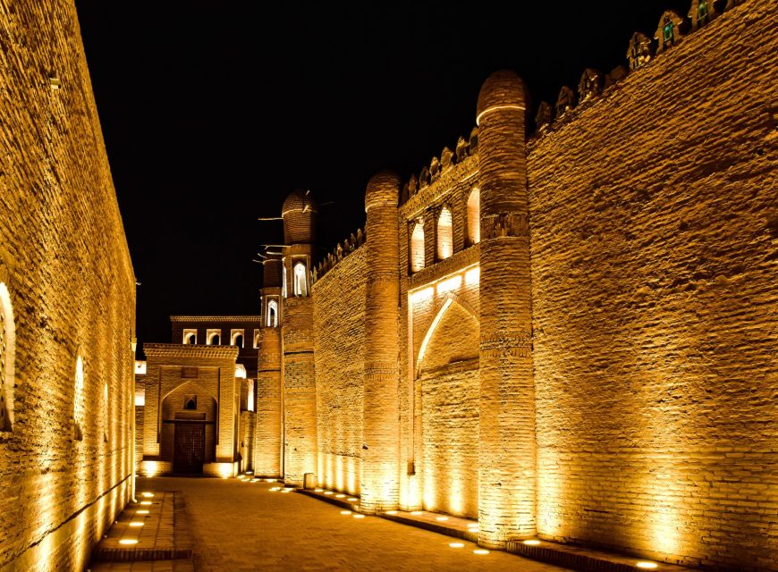 Night view of illuminated mud-brick fortress walls and cylindrical towers along a paved stone alleyway in the ancient historic city of Khiva, Uzbekistan.