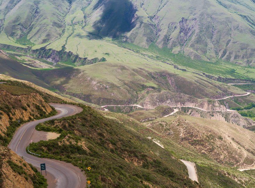 Elevated view of a narrow paved mountain road winding along steep, lush green hills in a alpine valley.