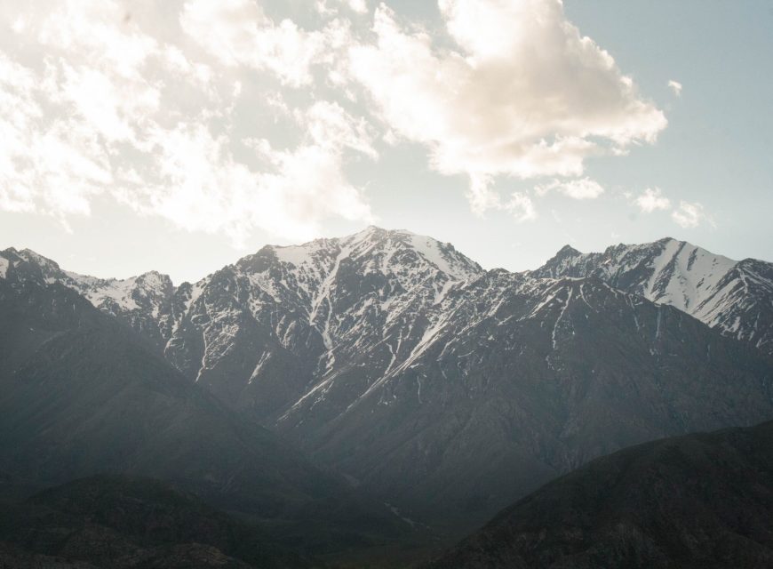 Black-and-white high-altitude landscape view of rugged mountain peaks reaching into a soft cloud-filled sky.