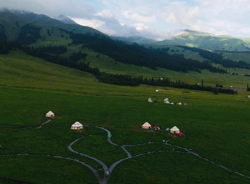 Aerial view of traditional white nomadic yurts scattered across a lush green alpine pasture with dirt pathways, set against misty, pine-covered mountain slopes under a soft sky.