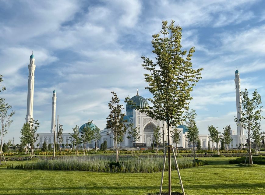 Low-angle view of the Astana Grand Mosque featuring white marble walls, blue-tilted domes, and towering minarets framed by a lush green park and bright blue sky with wispy clouds.